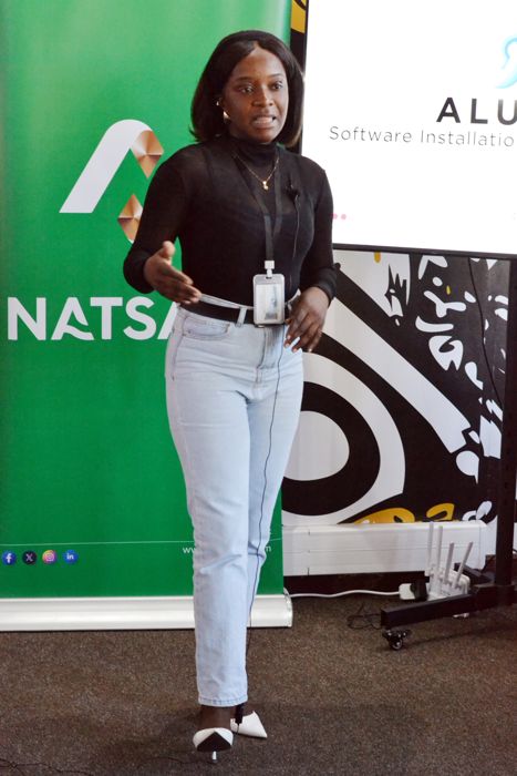 An Alusiny team member in a black top and light jeans speaks to the room, with a green NATSAVE banner beside her and a presentation on the screen behind.