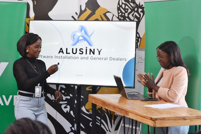 An Alusiny team member in a black top holds her microphone as the company logo and the line “Software Installation and General Dealers” show on screen, while a colleague applauds.