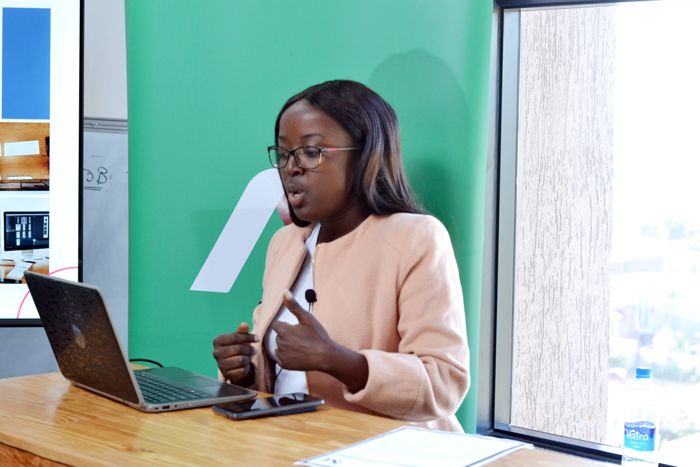 A colleague in a peach blazer explains a point from her laptop at the standing desk, with a green banner behind her.
