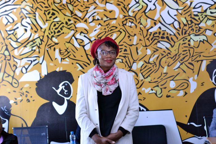 A guest in a red beret, floral scarf and cream coat smiles in front of the venue’s bold yellow and black mural.