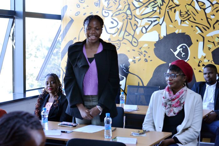 A participant in a black coat and lilac knit top stands to speak while a guest in a red beret and other attendees listen at the long table.