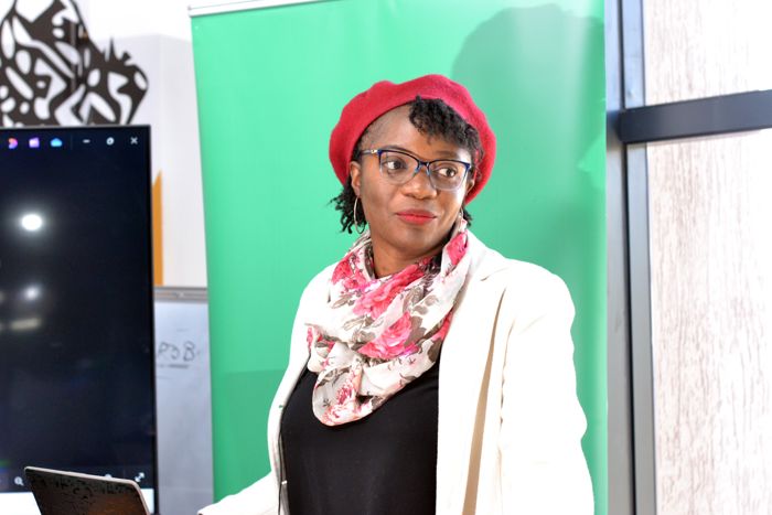 A guest in a red beret and glasses listens attentively, with a green banner and a screen behind her.