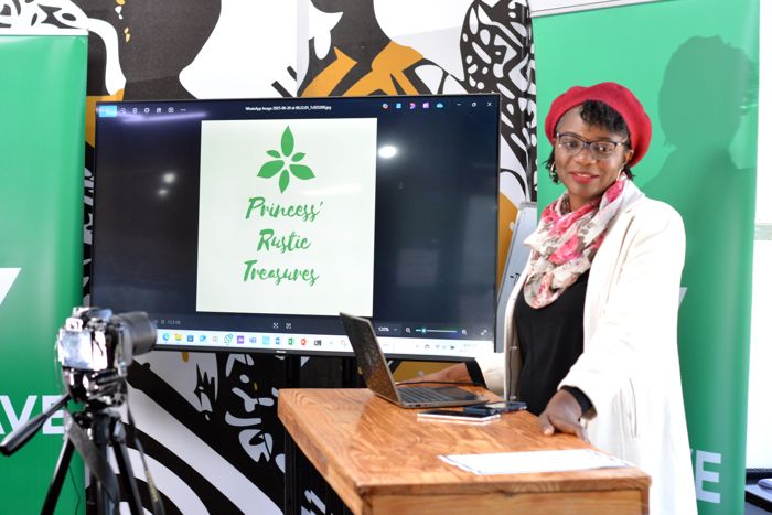 A guest in a red beret and floral scarf stands beside the screen showing the green leaf logo of “Princess’ Rustic Treasures”, an SME brand.