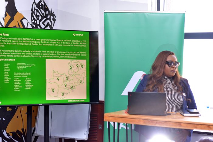 The speaker sits beside a screen showing a NATSAVE Bank slide about the bank’s history and its reach across Zambia’s provinces.