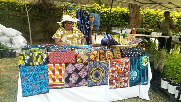 A stallholder in a yellow floral top sits behind neat stacks of brightly patterned African print fabrics.