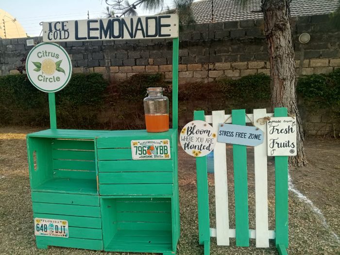 A green crate stand from “Citrus Circle” with a jar of fresh lemonade and signs reading “Bloom where you are planted” and “Stress free zone”.