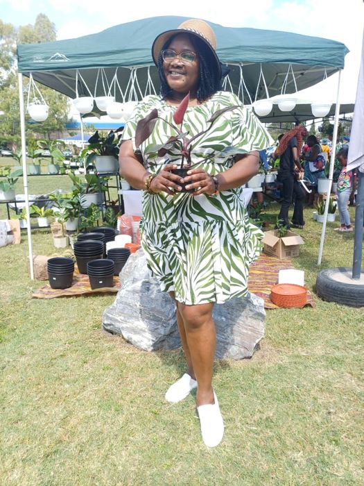 A smiling visitor in a straw hat and green leaf-print dress holds a red-leaved plant while standing on a rock in front of a teal canopy.