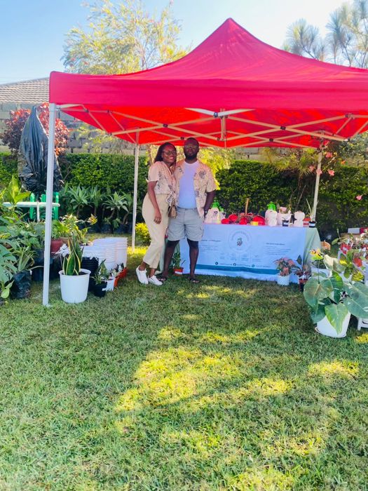 A couple in matching floral shirts pose together at their stall under a red canopy, with plants and pots around them.