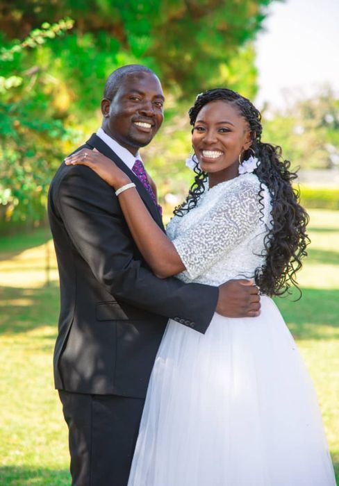 The couple embrace and smile on a sunlit lawn, the groom in a black suit with a purple tie and the bride in a beaded lace-sleeved gown.