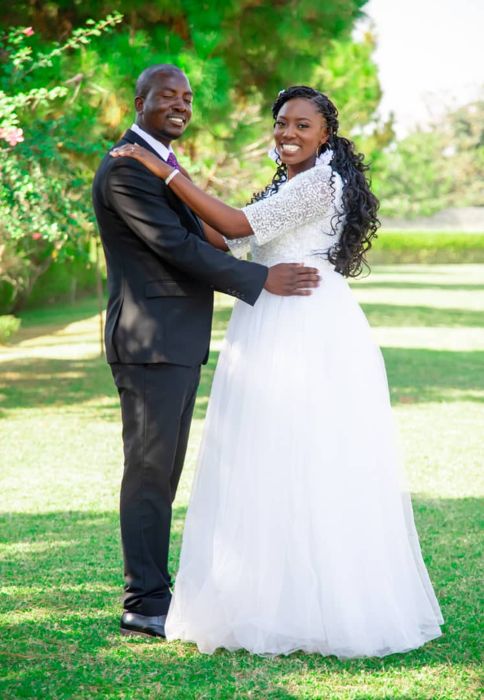 The bride rests her hand on the groom’s shoulder, her long curled hair over one shoulder, as they stand together on the grass.