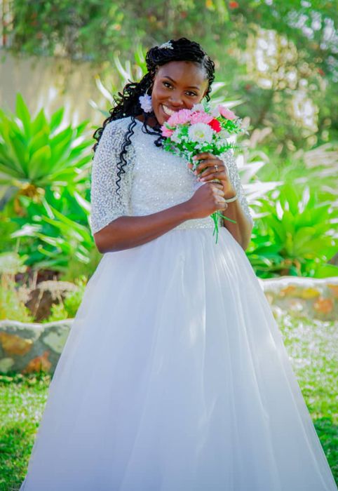 The bride smiles with a bouquet of pink and white flowers held to her cheek, green agave plants behind her.