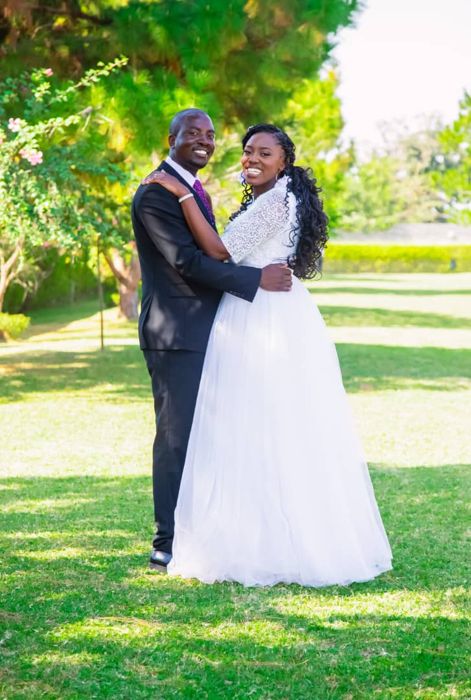 The couple smile at the camera in front of flowering bushes, the groom in a dark suit and the bride in a long tulle skirt.
