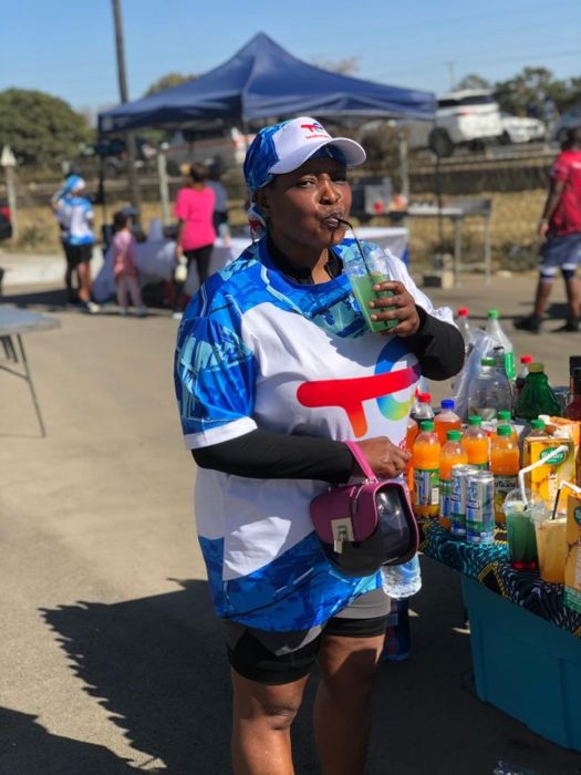 A participant in a blue TotalEnergies shirt and cap sips a drink through a straw beside a table of juices and soft drinks.