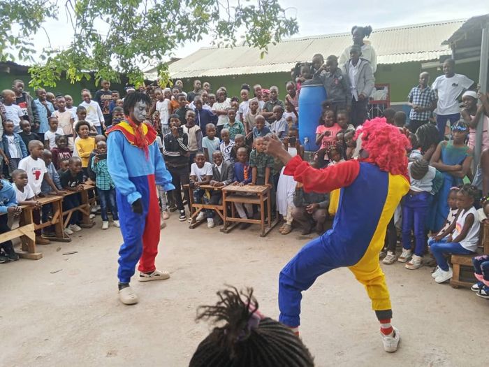 Two performers in colourful clown costumes entertain a large crowd of children gathered around them in a school yard.