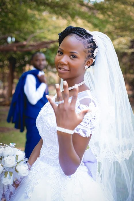 The bride holds up her hand to show her ring, framed by her veil and a bouquet of white roses, while the groom stands in a blue waistcoat behind her.