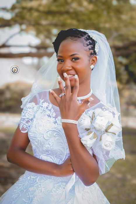 The bride in a lace gown and sparkling headpiece smiles as she shows her ring, with a bouquet of white roses at her shoulder.