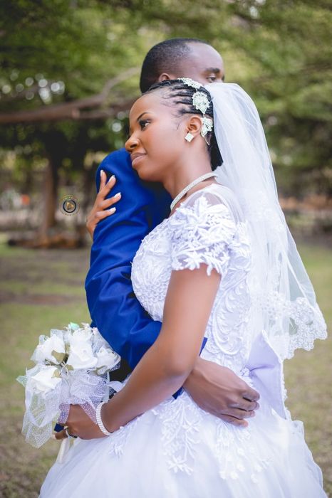 The couple embrace in profile under the trees, the bride in a lace-sleeved gown and veil with the groom’s blue jacket wrapped around her.