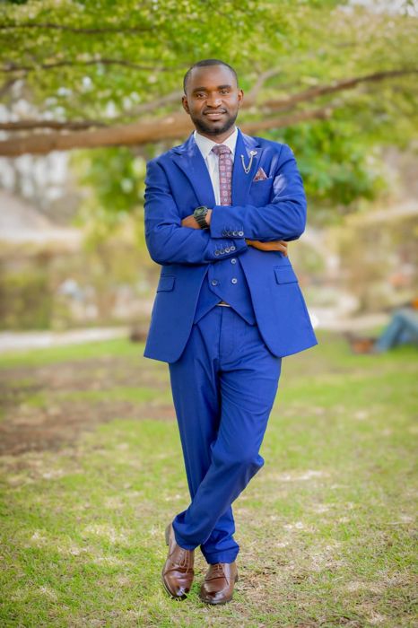 The groom in a bright blue three-piece suit and patterned tie stands with his arms crossed under a spreading tree.