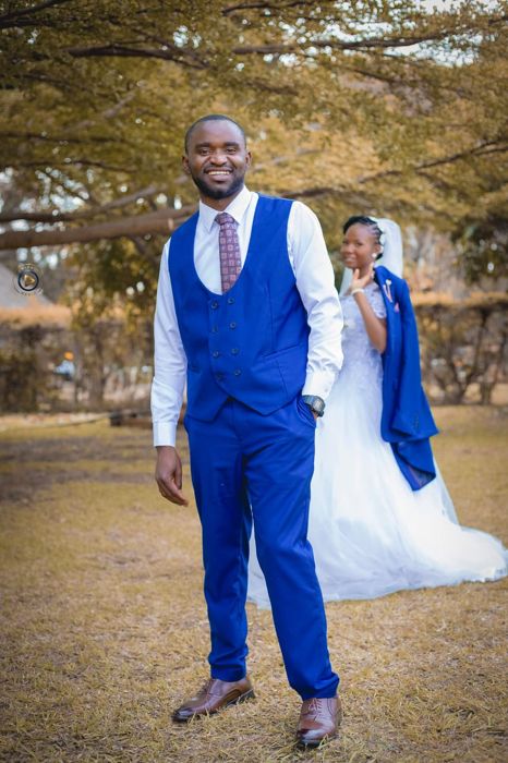 The groom smiles at the camera in a blue waistcoat while the bride, in her veil, peeks out behind him with his jacket over her arm.