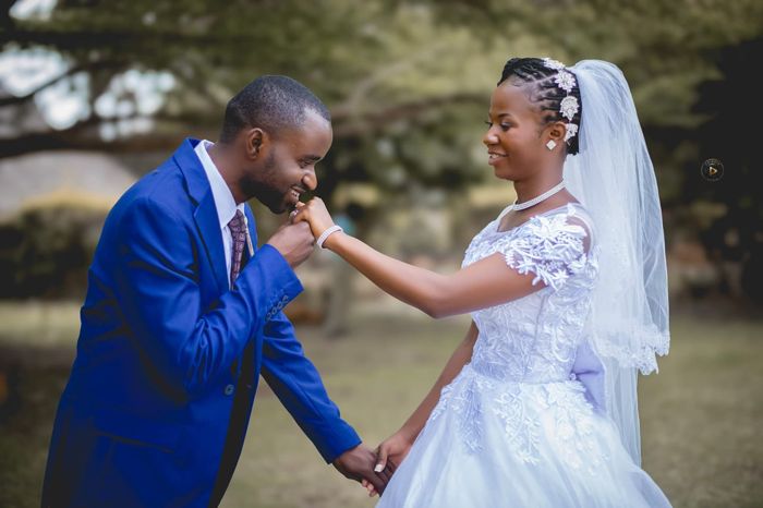The groom bends to kiss the bride’s hand as she smiles down at him, both framed by the trees.