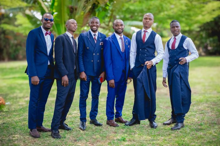 The groom, in bright blue, stands with five groomsmen in navy suits, waistcoats and bow ties, all smiling on the lawn.