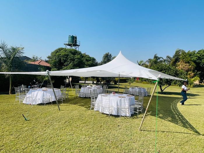 A white stretch tent shelters round tables laid with white cloths and clear chairs on a sunny garden lawn.