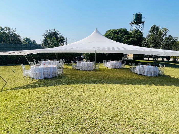Round tables dressed in white sit under the peaked white tent, with a green water tower beyond the trees.